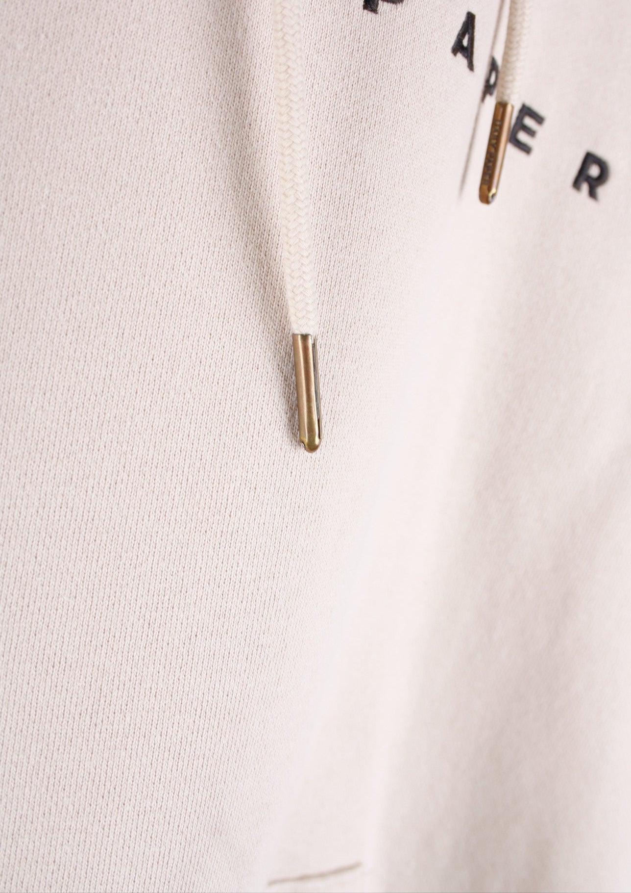 Close-up van de beige Daily Paper Alias hoodie met touwtjes en goudkleurige uiteinden, die een luxe en verfijnde afwerking bieden.