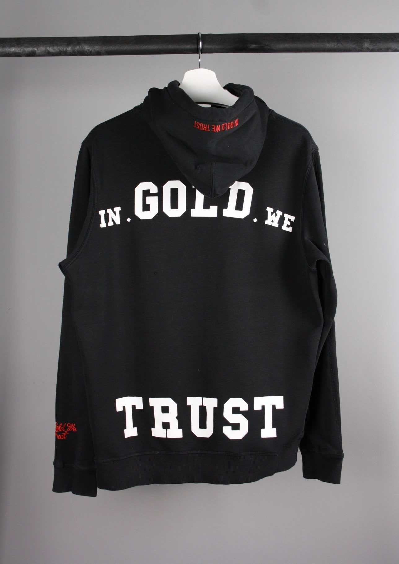 Achterkant van een zwarte 'In Gold We Trust' hoodie met een groot merklogo op de rug, perfect voor een trendy en casual outfit.