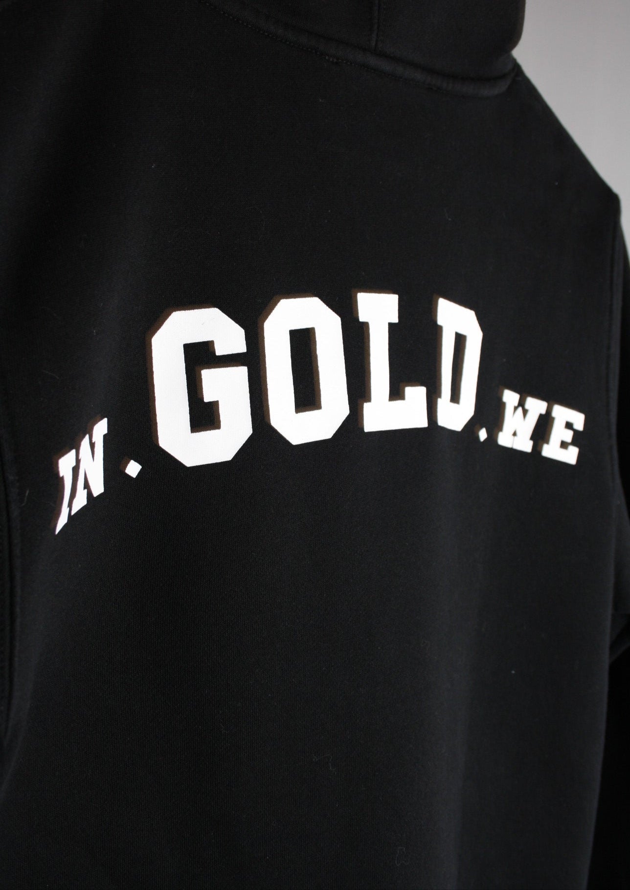 Close-up van het grote witte In Gold We Trust logo op de achterkant van een zwarte hoodie, met strakke print en premium afwerking voor een moderne streetwear uitstraling."