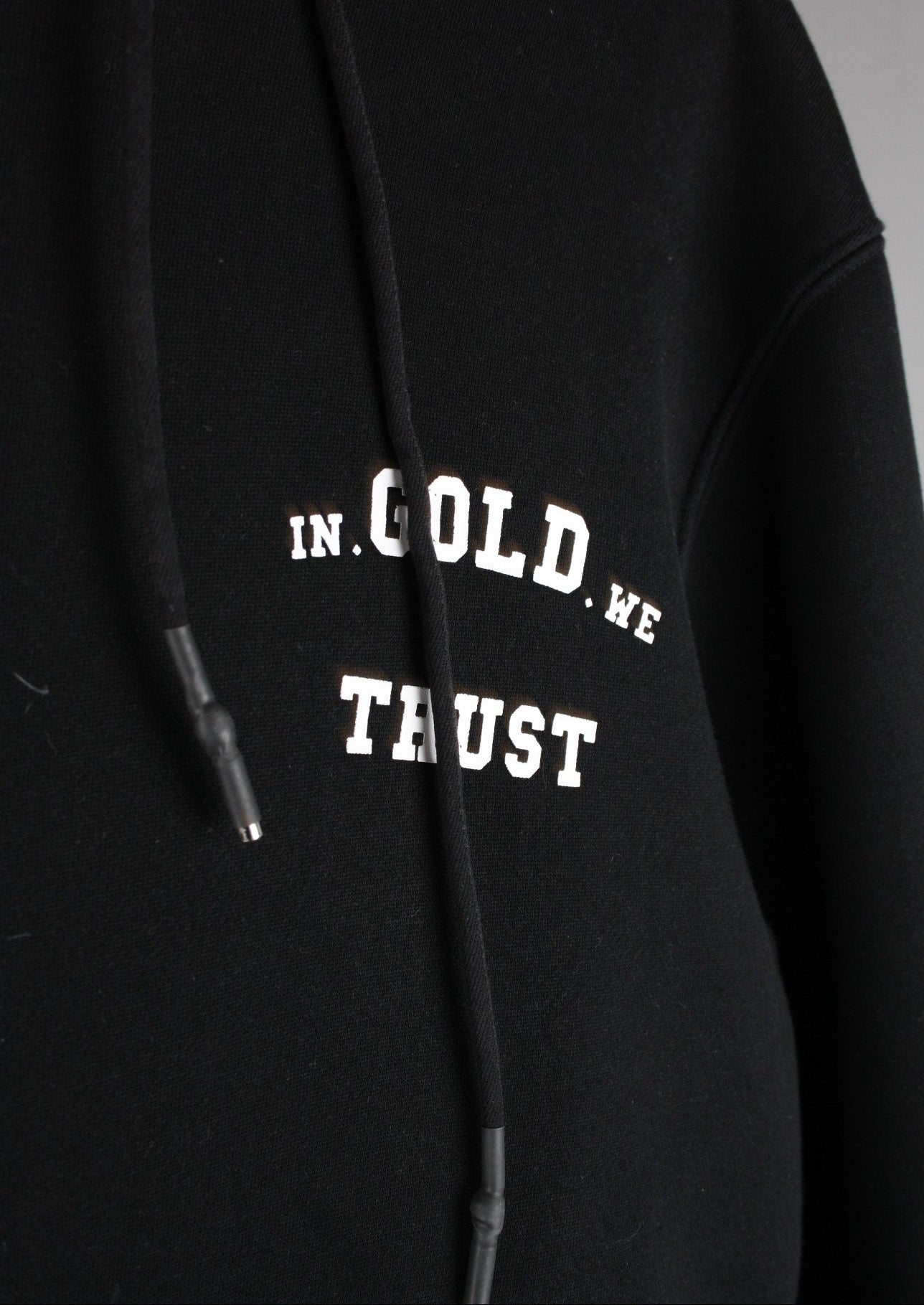 Close-up van het witte In Gold We Trust logo op de voorkant van een zwarte hoodie, met een strak gedrukt design voor een moderne streetwear look.
