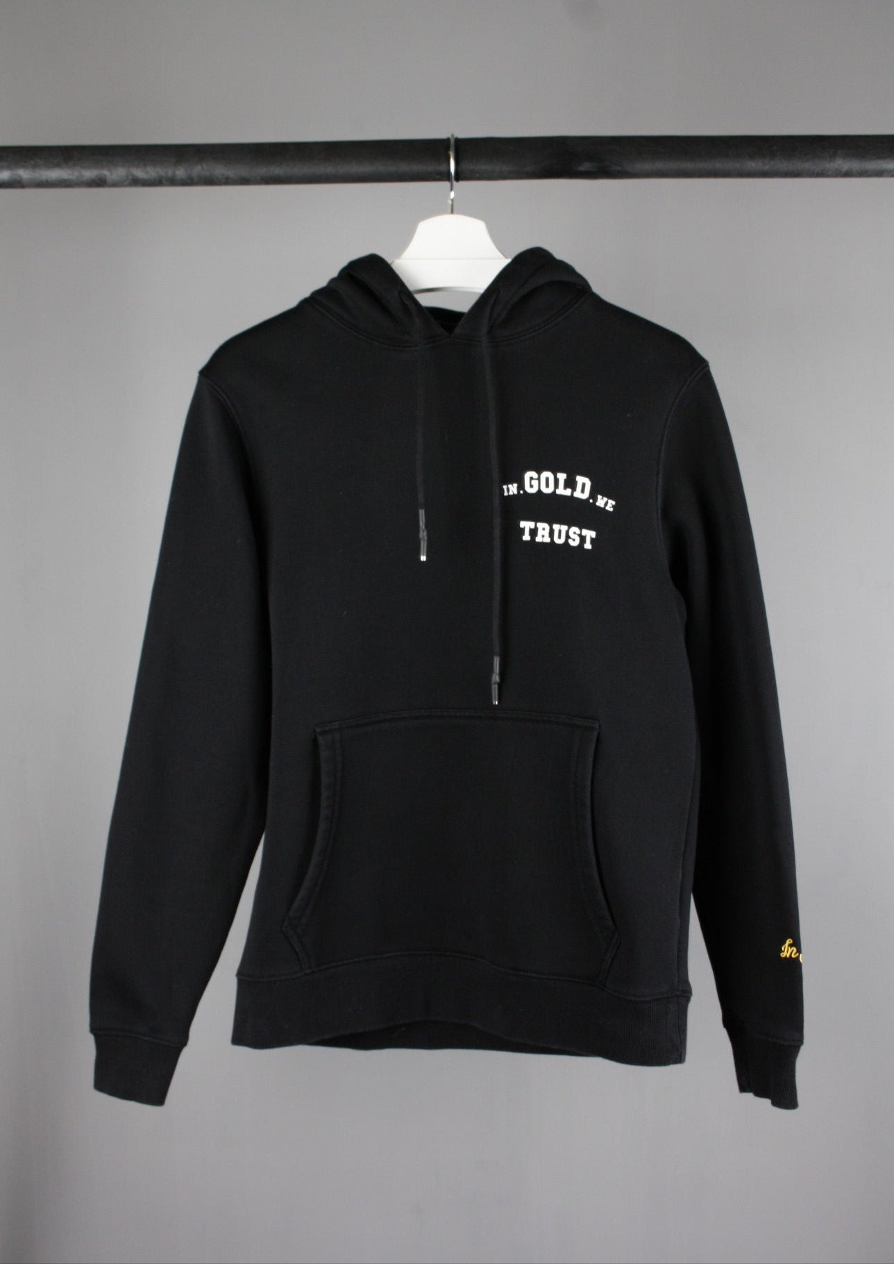 Voorkant van een zwarte In Gold We Trust hoodie met een wit logo op de borst, casual en streetwear geïnspireerd design.
