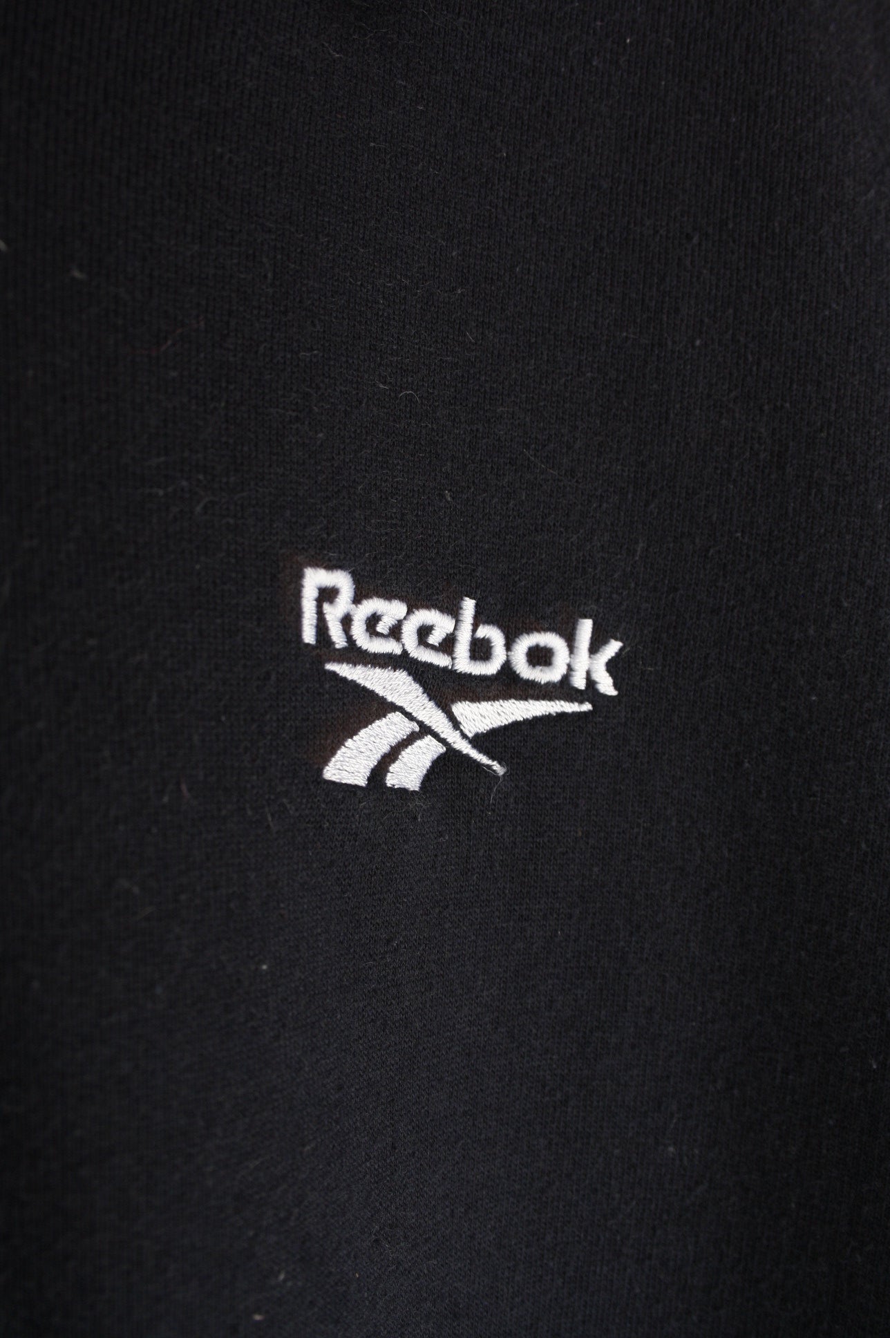 Close-up van het wit geborduurde logo op de voorkant van een marineblauwe Reebok quarter zip sweater in maat S, het merkiconische logo.