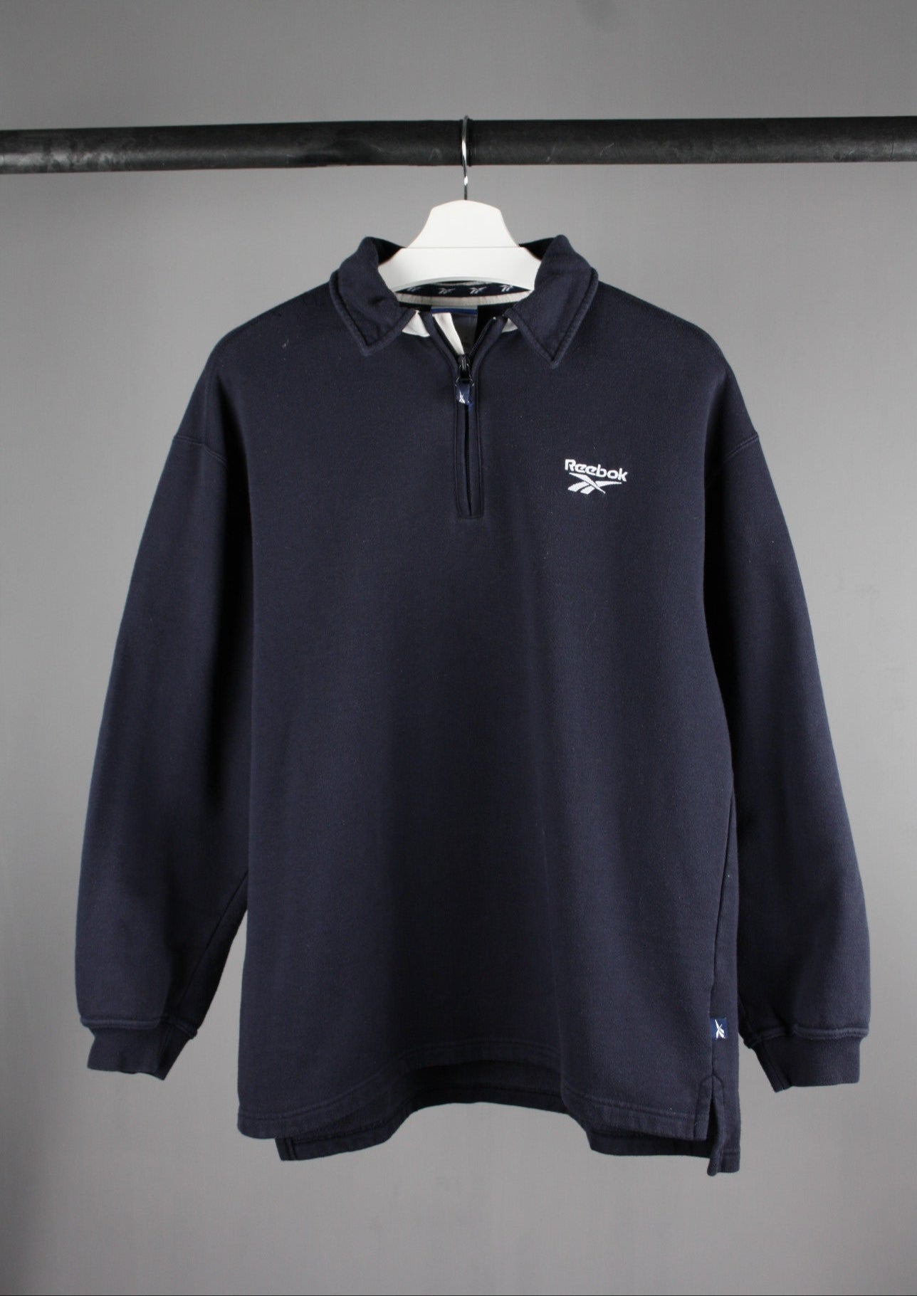 Voorkant van een marineblauwe Reebok quarter zip sweater in maat S met een wit geborduurd logo, voor een sportieve en casual uitstraling.