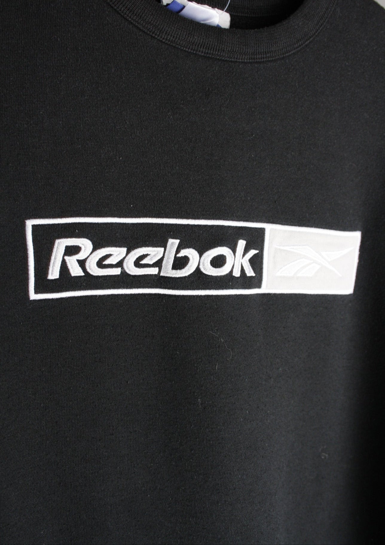 Close-up van het grote geborduurde logo op de borst van een zwarte Reebok sweater in maat XS.
