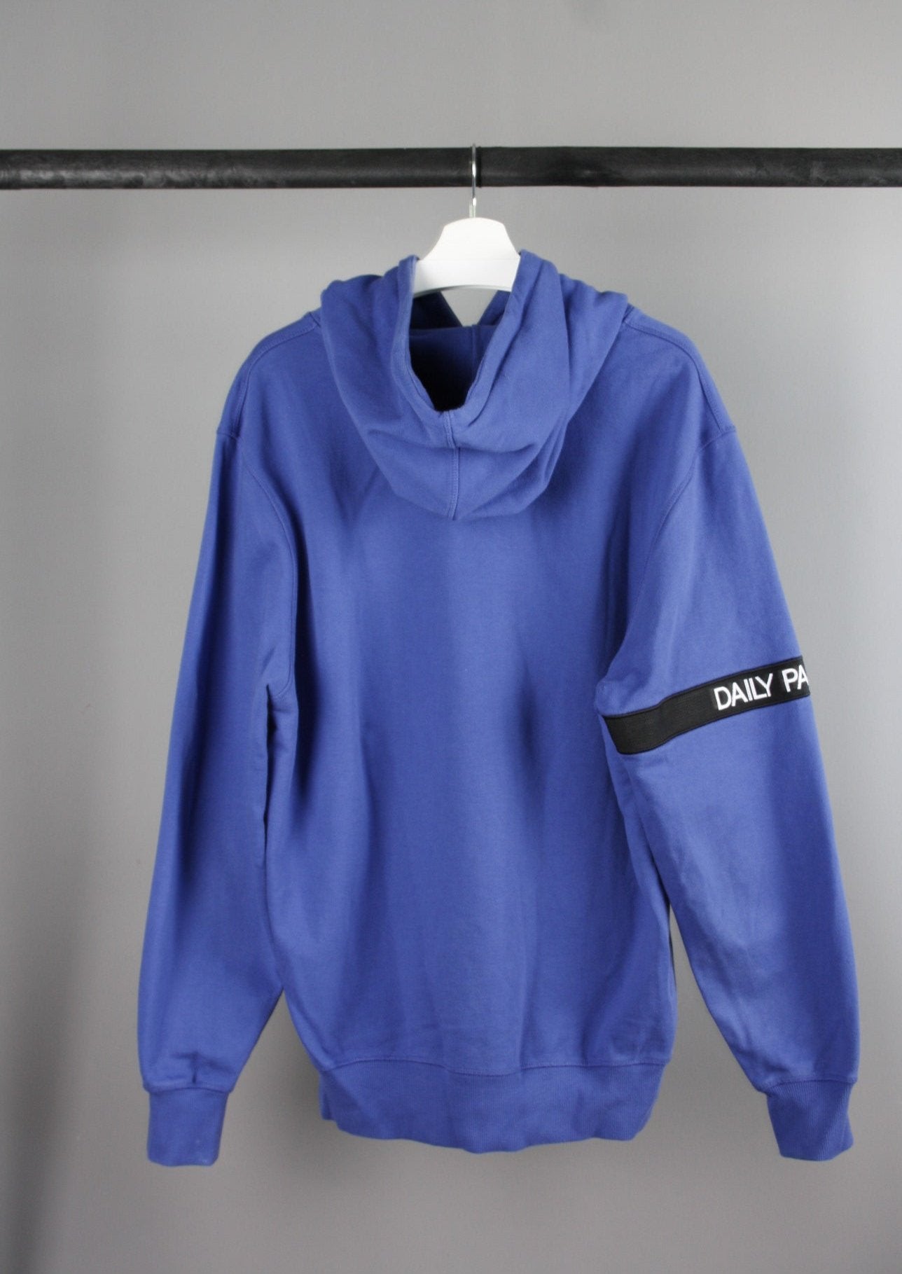 Achterkant van een blauwe Daily Paper Captain hoodie met een zwarte band op de rechterarm, ideaal voor een moderne en stijlvolle look.
