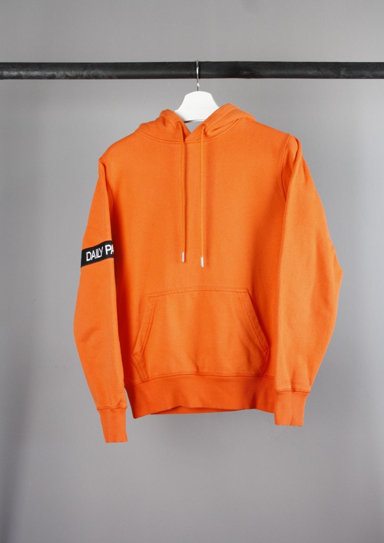 Voorkant van een oranje Daily Paper Captain hoodie met een zwarte band om de rechterarm, gemaakt van comfortabele stof met een opvallende kleurcombinatie.