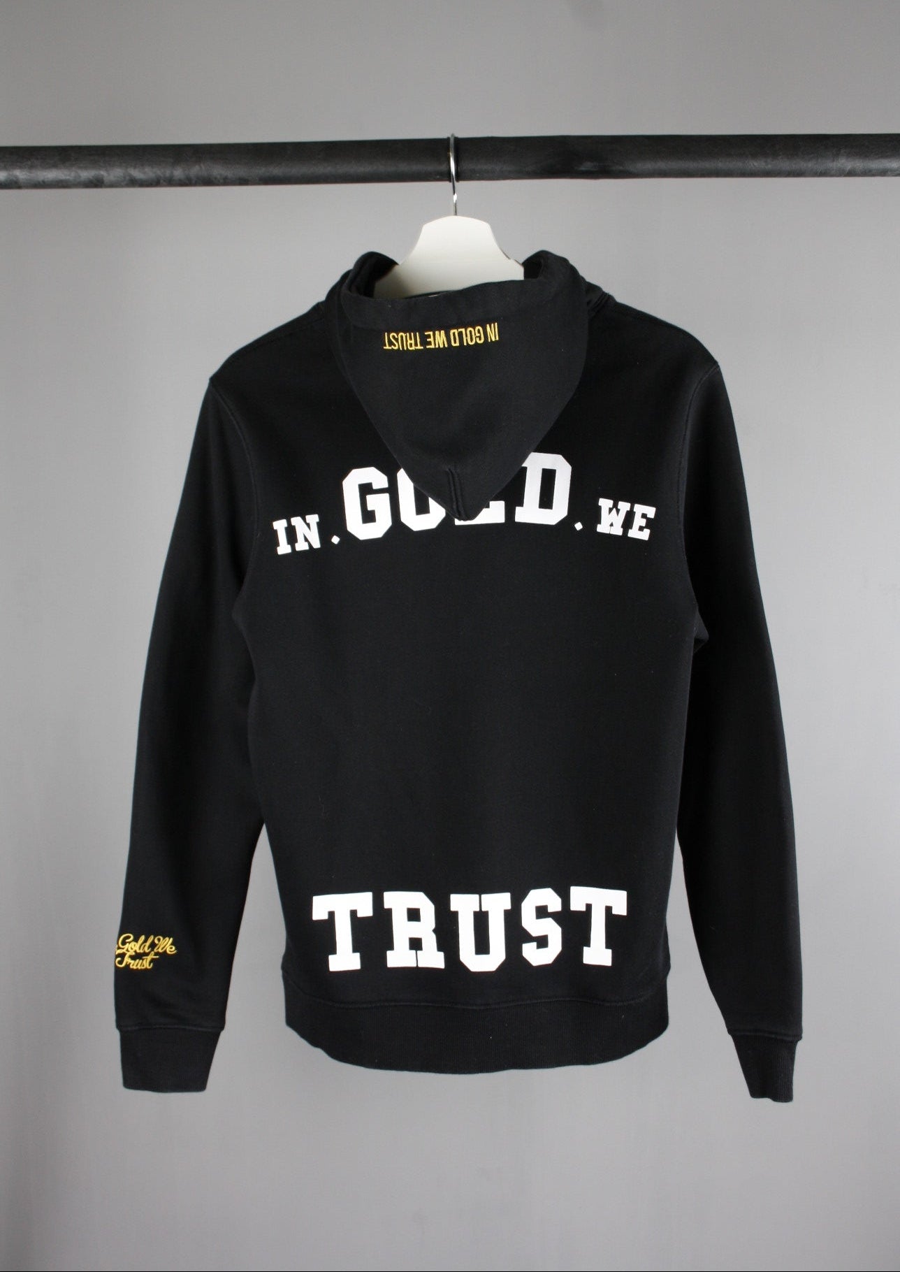 Achterkant van een zwarte In Gold We Trust hoodie met groot wit logo, ideaal voor een trendy en urban look.