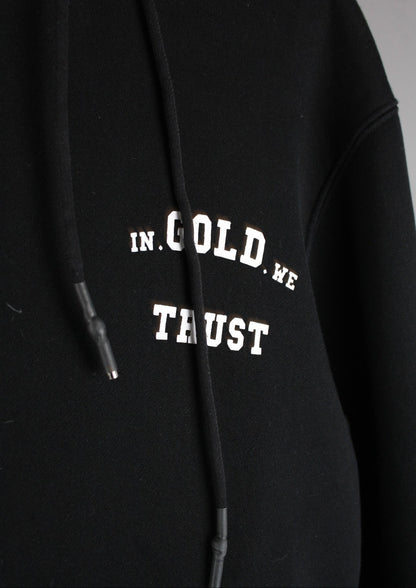 Close-up van het witte In Gold We Trust logo op de voorkant van een zwarte hoodie, met een strak gedrukt design voor een moderne streetwear look.