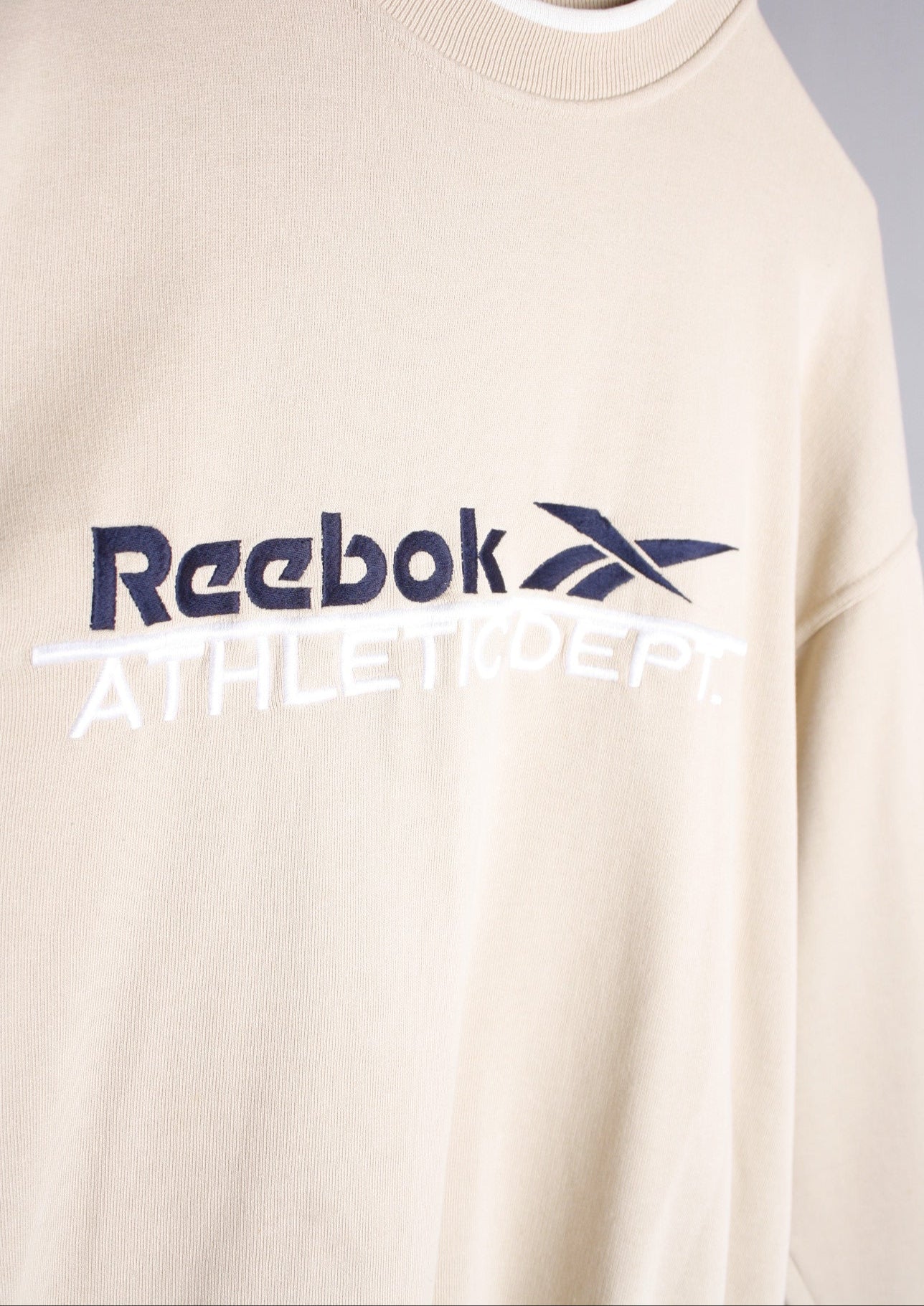 Close-up van het groot marineblauw geborduurde logo op de borst van een beige Reebok Athletic Dept sweater in maat S.
