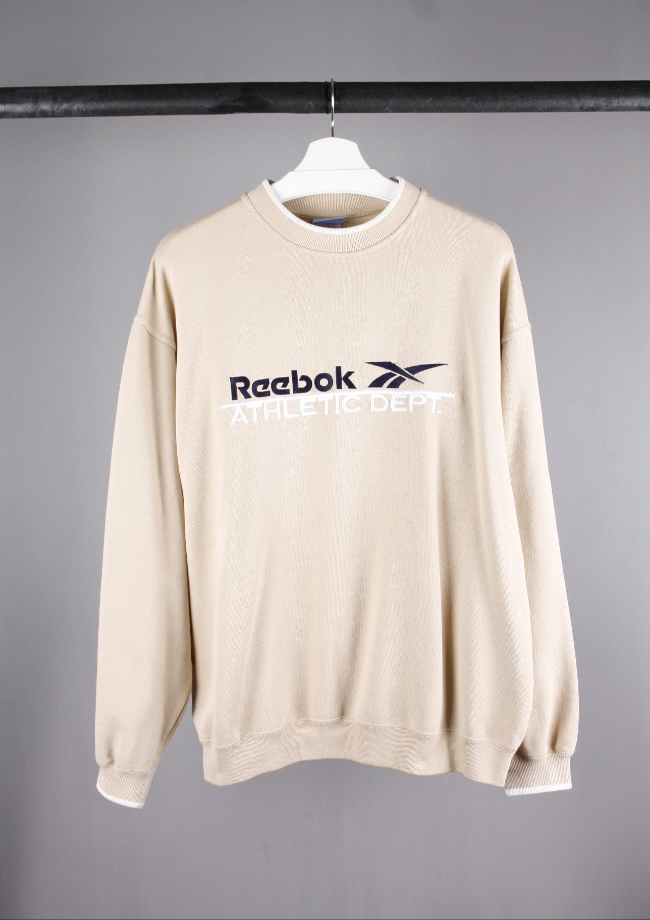 Voorkant van een beige Reebok Athletic Dept sweater in maat S met een groot marineblauw geborduurd logo op de borst, ideaal voor een sportieve en casual look.