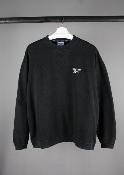 Voorkant van een zwarte Reebok sweater in maat L met een wit geborduurd logo linksboven, voor een sportieve en casual uitstraling.